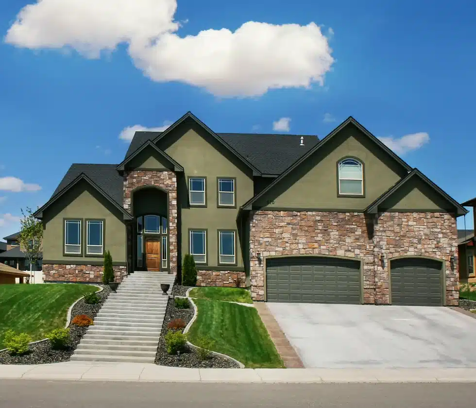 A large two-story home with olive green stucco, stone veneer accents, a three-car garage, and a wide staircase leading to the front entry.